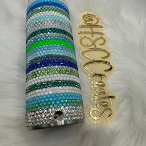 May include: A stainless steel tumbler with a colorful design. The tumbler is covered in rhinestones in shades of blue, green, and white. The tumbler is decorated with a striped pattern.