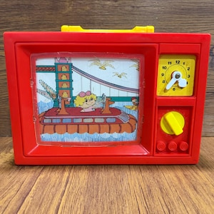 May include: A vintage red plastic toy television with a yellow handle and dial. The screen displays a cartoon scene of a bridge and characters. The clock on the side has a white clock face with black numbers and hands. The toy is on a wooden surface.