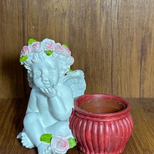 May include: A white cherub figurine with a floral crown and a red planter. The cherub is seated, with its hand near its face. The planter is a small, round pot with vertical ridges. The cherub has wings and is decorated with pink roses.