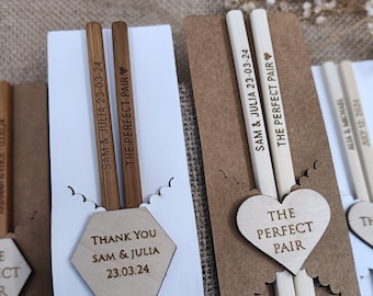 Könnte beinhalten: Zwei Sätze Holzstäbchen mit personalisierten Gravuren. Die Stäbchen sind in braunes Papier verpackt mit einem weißen herzförmigen Etikett, auf dem "The Perfect Pair" steht.