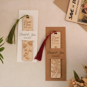 Könnte beinhalten: Zwei Holz-Lesezeichen mit einem floralen Design und dem Text "David & Jane" und dem Datum "04.05.2025". Die Lesezeichen sind in weißen und braunen Umschlägen mit roten und grünen Quasten verpackt.