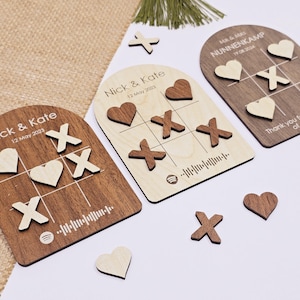 Op de afbeelding: Twee houten tic-tac-toe borden met gepersonaliseerde namen en data. De borden zijn boogvormig en hebben een bruine houten afwerking. De spelstukken zijn harten en X'en.