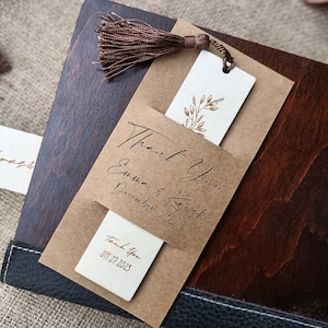 Pode incluir: Um marcador de p&aacute;gina de papel castanho com um inserto branco com um design floral dourado. O marcador de p&aacute;gina tem uma borla castanha e o texto "Thank you Emma & Krish December 2023" e "Thank You 01.12.2023".