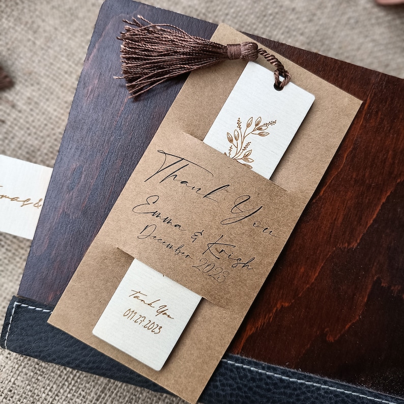 Pode incluir: Um marcador de p&aacute;gina de papel castanho com um inserto branco com um design floral dourado. O marcador de p&aacute;gina tem uma borla castanha e o texto "Thank you Emma & Krish December 2023" e "Thank You 01.12.2023".
