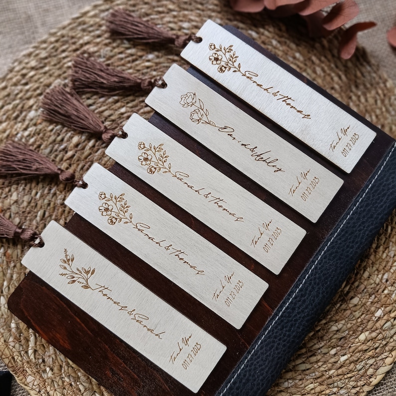 Pode incluir: Conjunto de seis marcadores de p&aacute;gina de madeira com um design floral e o texto "Sarah & Thomas" e "Thomas & Sarah" com uma data. Cada marcador de p&aacute;gina tem uma borla marrom.