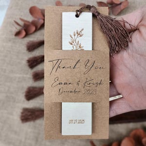 Pode incluir: Um marcador de p&aacute;gina de papel castanho com um marcador de p&aacute;gina de madeira branca no seu interior. O marcador de p&aacute;gina de madeira tem um desenho floral dourado e o texto "Thank You Emma & Krish December 2023 01.27.2023".