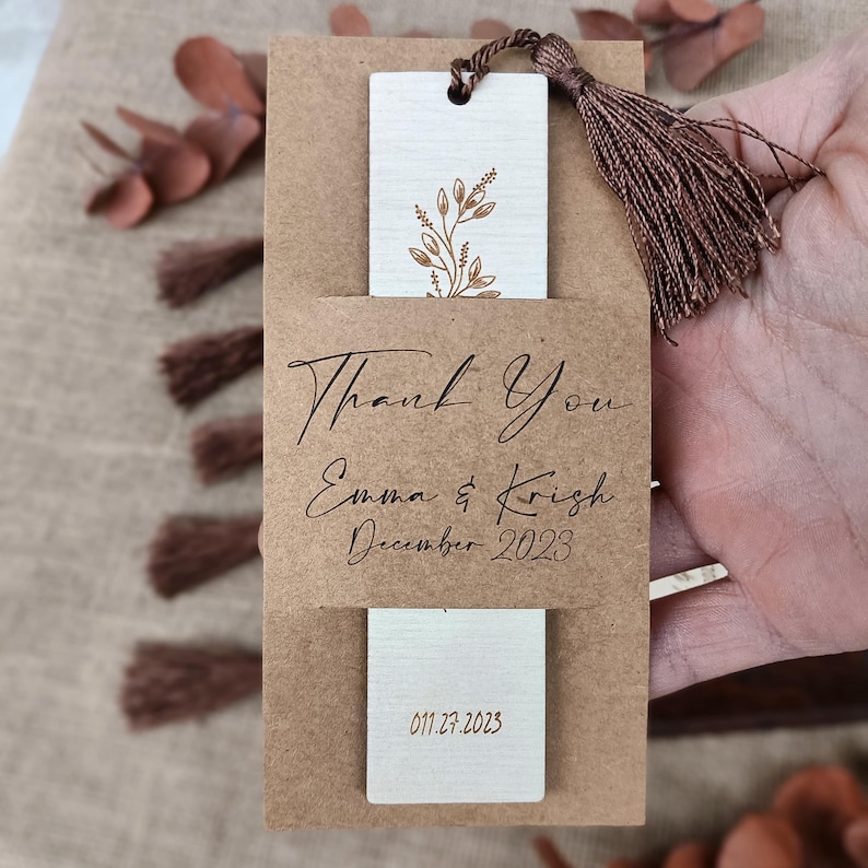Pode incluir: Um marcador de p&aacute;gina de papel castanho com um marcador de p&aacute;gina de madeira branca no seu interior. O marcador de p&aacute;gina de madeira tem um desenho floral dourado e o texto "Thank You Emma & Krish December 2023 01.27.2023".