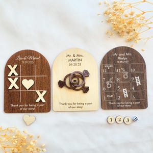 Könnte beinhalten: Drei Hochzeitsgeschenke aus Holz. Eines ist ein Tic-Tac-Toe-Spiel, ein anderes zeigt ineinandergreifende Ringe und das dritte ist ein Brettspiel mit Leitern und Schlangen. Jedes hat den Namen und das Hochzeitsdatum eines Paares. Enthält Spielsteine aus Holz.