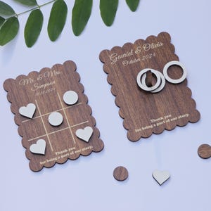 Könnte beinhalten: Zwei Hochzeitsspiele aus Holz. Eines ist ein Tic-Tac-Toe-Spiel mit weißen Herz- und Kreisstücken. Das andere ist ein Ringwurfspiel mit zwei weißen Ringen. Beide Spiele haben eine personalisierte Nachricht mit den Namen des Paares und dem Hochzeitsdatum. "Mr. & Mrs. Simpson 10.13.2024" und "Gavriel & Olivia October 2024". Beide Spiele haben die Nachricht "Vielen Dank, dass Sie Teil unserer Geschichte sind."
