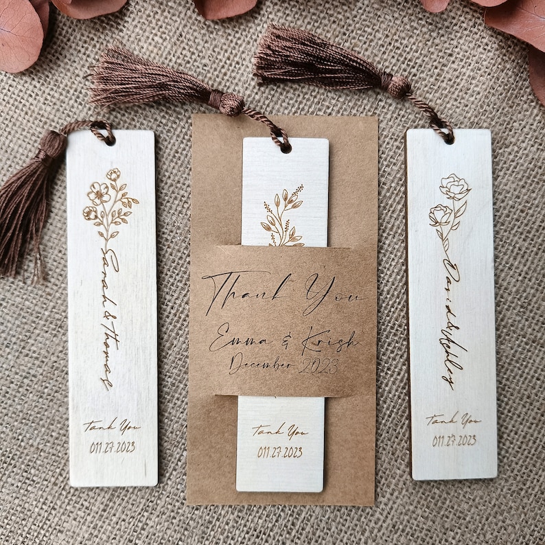 Pode incluir: Tr&ecirc;s marcadores de p&aacute;gina de madeira com borlas marrons. Cada marcador de p&aacute;gina apresenta um design floral e o texto "Obrigado" com uma data. O marcador de p&aacute;gina do meio tamb&eacute;m inclui o texto "Emma & Krish Dezembro 2023".