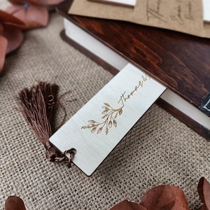 Pode incluir: Um marcador de p&aacute;gina de madeira com um design floral dourado e a palavra "Thomas" gravada nele. O marcador de p&aacute;gina tem uma borla marrom.