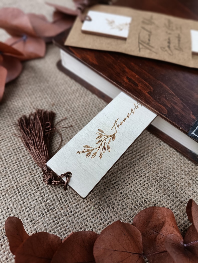 Pode incluir: Um marcador de p&aacute;gina de madeira com um design floral dourado e a palavra "Thomas" gravada nele. O marcador de p&aacute;gina tem uma borla marrom.