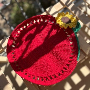 May include: A handmade, red crochet placemat with a decorative sunflower accent. The placemat is round, with a scalloped edge. The sunflower has yellow petals and a brown center, with green leaves and stem.