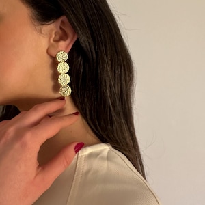 Peut inclure: Boucles d'oreilles pendantes dorées avec un motif martelé texturé. Chaque boucle d'oreille comporte quatre disques circulaires suspendus verticalement. Les boucles d'oreilles sont portées par une personne aux cheveux foncés et un haut blanc.