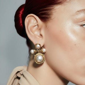May include: A gold-toned earring featuring a central large white pearl, accented by three smaller white pearls arranged in a cross-like structure. The metal setting has a textured, circular frame around the main pearl and a polished finish.