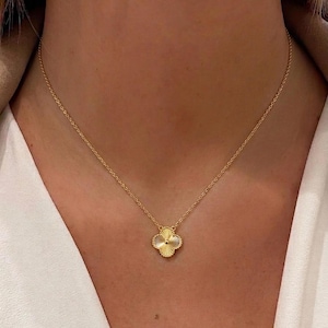 Op de afbeelding: Een goudkleurige ketting met een vierbladig klaverblad hanger. De hanger heeft een getextureerd, geribbeld ontwerp met een klein, rond midden. De ketting is delicaat en lijkt van goud te zijn. De ketting wordt om de nek gedragen.
