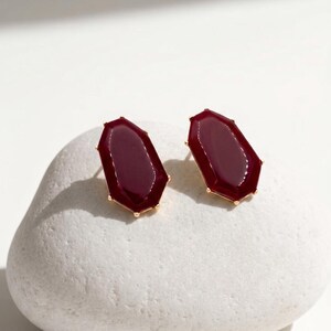 May include: A pair of stud earrings featuring a deep burgundy, geometric-shaped stone set in a gold-colored metal frame. The earrings are displayed on a white, textured surface, showcasing their polished finish and design.