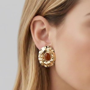 May include: Gold-coloured stud earrings with a textured surface and a central oval amber-coloured stone. The earrings have a hammered metal appearance. The earrings are a statement piece and are worn on the ear.