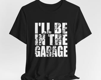 May include: Black t-shirt with white text that reads "I'll be in the garage". The text has a distressed, tire tread-like pattern.
