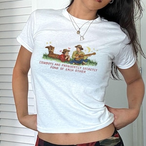 May include: White t-shirt with a graphic of three cowboys on a grassy hill, one playing a guitar. The text "Cowboys are frequently secretly fond of each other" is printed below the graphic. The shirt is short-sleeved.