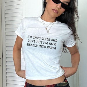 May include: White t-shirt with the text "I'M INTO GIRLS AND GUYS BUT I'M ALSO REALLY INTO PASTA" in black. The person is wearing black sunglasses and a floral skirt with red and black flowers.