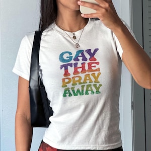 Peut inclure: T-shirt blanc avec l'inscription "GAY THE PRAY AWAY" aux couleurs de l'arc-en-ciel. Le t-shirt a un col rond et des manches courtes. Le texte est en gras et scintillant.