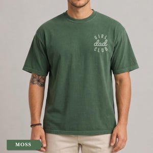 Puede incluir: Camiseta verde musgo con el texto "GIRL dad CLUB" en escritura blanca en la parte superior derecha del pecho. La camiseta tiene cuello redondo y mangas cortas. La camiseta es de color liso y tiene un aspecto ligeramente descolorido.