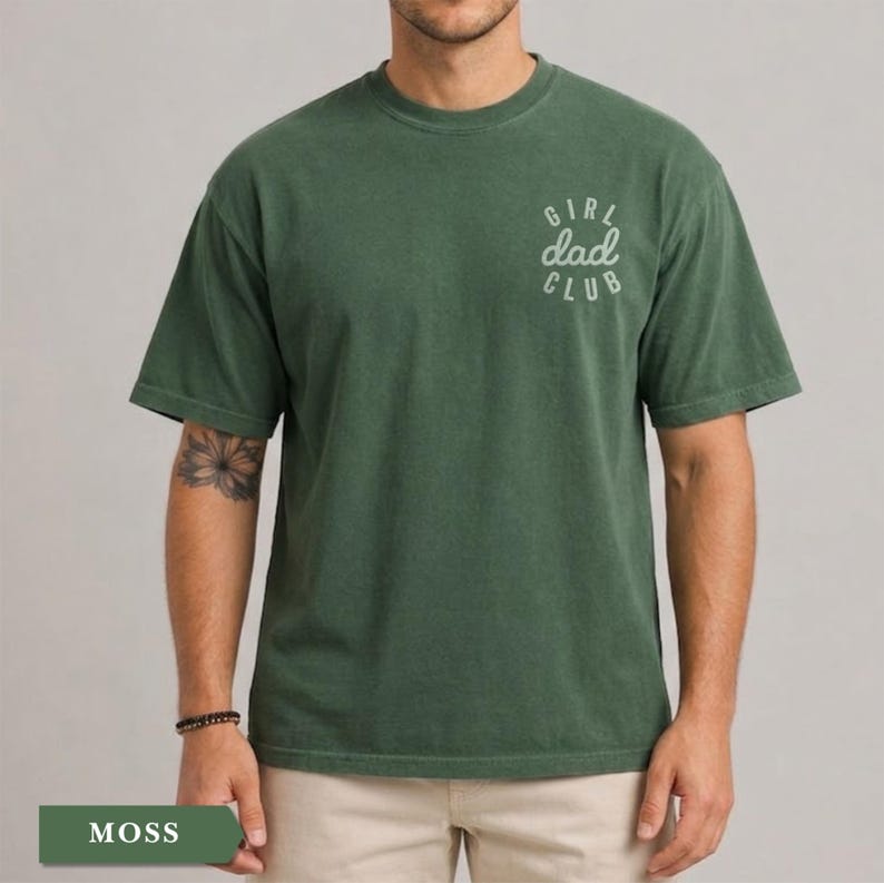 Puede incluir: Camiseta verde musgo con el texto "GIRL dad CLUB" en escritura blanca en la parte superior derecha del pecho. La camiseta tiene cuello redondo y mangas cortas. La camiseta es de color liso y tiene un aspecto ligeramente descolorido.
