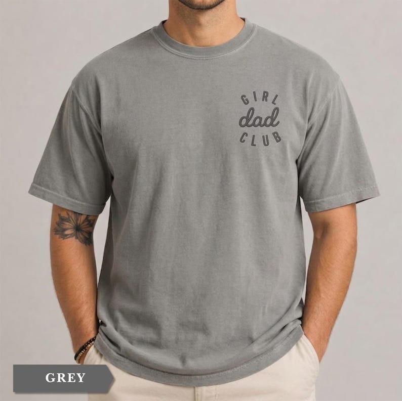 Puede incluir: Camiseta gris con mangas cortas. La camiseta tiene la frase "GIRL dad CLUB" impresa en la parte superior derecha del pecho. La camiseta es de un material suave y tiene un corte relajado.