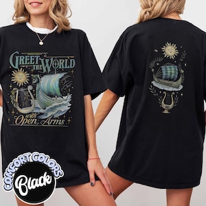May include: Black t-shirt with a front graphic featuring a Viking ship sailing on waves, a sun, and the text "Greet the World with Open Arms." The back of the shirt has a similar design. The shirt is labeled "Comfort Colors Black."