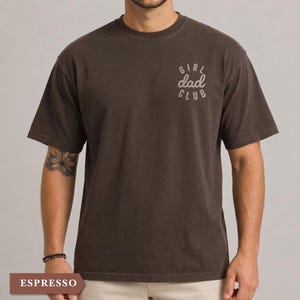 Puede incluir: Camiseta color espresso con mangas cortas. La camiseta presenta el texto "GIRL dad CLUB" en gris claro en el pecho. La camiseta es de color liso y tiene un corte holgado.