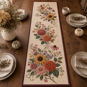 Può includere: Un runner da tavola trapuntato rettangolare con sfondo color crema e un intricato motivo floreale di girasoli, zinnie e margherite nei toni dell'arancione, giallo, rosa e viola, rifinito con un bordo di colore viola scuro.