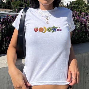 May include: White t-shirt featuring an embroidered fruit design. The design includes a strawberry, orange slice, banana, kiwi slice, blueberries, and cherries. The shirt has a crew neck and short sleeves.