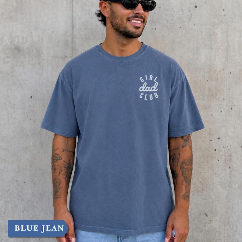 Puede incluir: Camiseta azul vaquero de manga corta con el texto "GIRL DAD CLUB" en escritura blanca en la parte superior de la espalda. La camiseta es de color azul liso y tiene un corte holgado. La parte inferior de la imagen tiene el texto "BLUE JEAN".