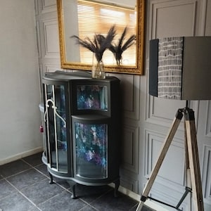 May include: A black display cabinet with glass doors and shelves. The cabinet has a curved front and sits on four legs. A vase with black dried flowers sits on top of the cabinet. A black and white patterned lamp with a wooden tripod stand is in the background.