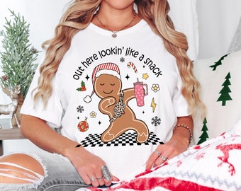 May include: White t-shirt with a Christmas-themed graphic. The design features a gingerbread man wearing a Santa hat and a leopard print fanny pack, holding a pink tumbler. Text reads "out here lookin' like a snack."