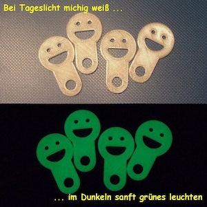 Könnte beinhalten: Fünf kleine, runde, weiße Schlüsselanhänger mit einem Smiley-Gesicht. Die Schlüsselanhänger leuchten im Dunkeln grün.