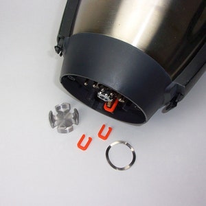 Könnte beinhalten: Ein zerlegter Mixer-Sockel mit einem silbernen Metallgehäuse, einem schwarzen Plastikdeckel und einem silbernen Metallring. Außerdem gibt es drei kleine rote Plastikclips und ein silbernes Metallteil mit sternförmigem Design.