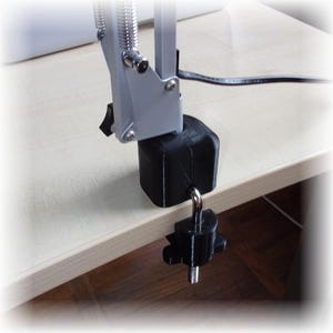 Könnte beinhalten: Eine silberne und schwarze Schreibtischlampenklemme, befestigt an einer hellfarbigen Holzoberfläche. Die Klemme besteht aus schwarzem Kunststoff und verfügt über einen Schraubmechanismus zur Befestigung am Schreibtisch. Der Lampenarm ist silberfarben und hat einen Federmechanismus.