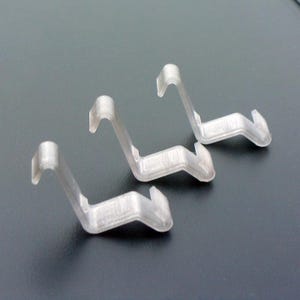 Könnte beinhalten: Drei transparente Kunststoffhaken, jeder mit einer gebogenen Form und einem spitzen Ende. Die Haken sind in einer Reihe angeordnet, wobei die spitzen Enden in die gleiche Richtung zeigen.