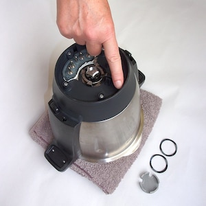 Könnte beinhalten: Ein zerlegter Mixer-Sockel mit einem silbernen Metallbehälter und einem schwarzen Plastikdeckel. Der Deckel hat eine Reihe von elektrischen Kontakten und ein kleines Metallzahnrad. Eine Hand drückt auf die Oberseite des Sockels. Drei kleine silberne Metallringe liegen auf einer grauen Oberfläche.