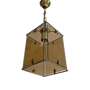 Könnte beinhalten: Vintage Pendelleuchte aus Messing und Glas. Die Leuchte hat eine geometrische, trapezförmige Form mit bernsteinfarbenen Glaspaneelen und einem Messingrahmen. Sie hängt an einer Kette und hat eine einzelne Glühbirne.