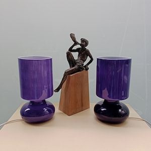 Könnte beinhalten: Zwei violette Tischlampen aus Glas mit rundem Sockel und zylindrischem Schirm. Zwischen den Lampen befindet sich eine Bronzeskulptur einer Person, die ein Horn spielt. Die Lampen sind an eine Stromquelle angeschlossen.