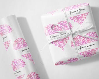 May include: White gift wrap and wrapping paper rolls with a pink floral heart design. The gift is tied with a white ribbon. The text "Joanne • Simon 14.06.25" is printed on the gift wrap.
