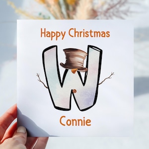 May include: A festive Christmas card featuring the words "Happy Christmas" in orange. The letter "W" is creatively designed as a snowman, complete with a brown top hat. The name "Connie" is written below.