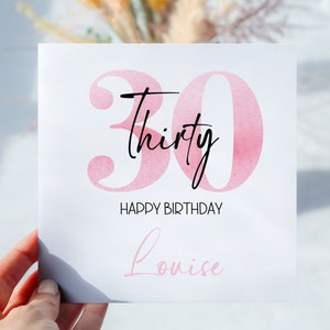 以下が含まれることがあります： ピンクの水彩画の誕生日カード。30という数字と「thirty」という単語が黒い筆記体で書かれています。カードには「Happy Birthday」と書かれており、「Louise」という名前でパーソナライズされています。