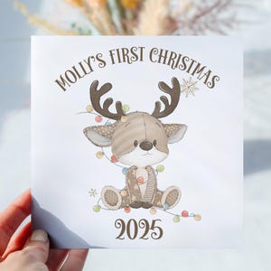 May include: A square Christmas card with the text "Molly's First Christmas" and the year "2025". The card features a cartoon reindeer with brown antlers, a light brown body, and a string of colourful lights.
