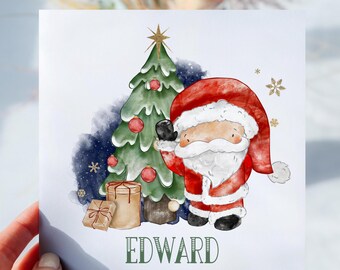May include: A personalised Christmas card featuring a cartoon Santa Claus holding a gift box next to a decorated Christmas tree. The card is white with a blue and green watercolour background and the name "EDWARD" printed in green at the bottom.
