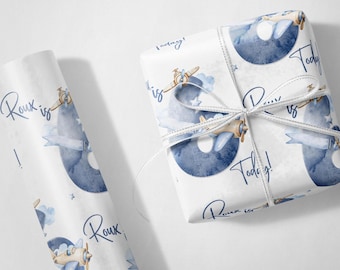 May include: White gift wrap with a blue watercolor design featuring a number 8, airplanes, clouds, and stars. The text "Roux is! Today!" is printed on the paper. A wrapped gift is tied with a white ribbon.