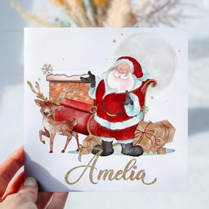 May include: A white card featuring a watercolour illustration of Santa Claus in a red suit with a white beard, delivering presents in a sleigh pulled by a reindeer. The card has a full moon in the background and the name "Amelia" written in gold glitter.